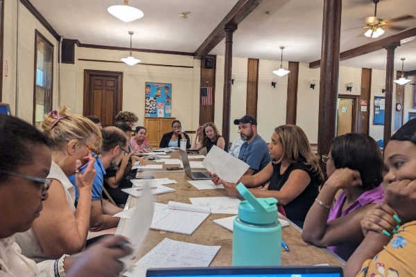 a committee meeting of people working to support refugee families in Woburn. About a dozen people seated around a long rectangle table. 