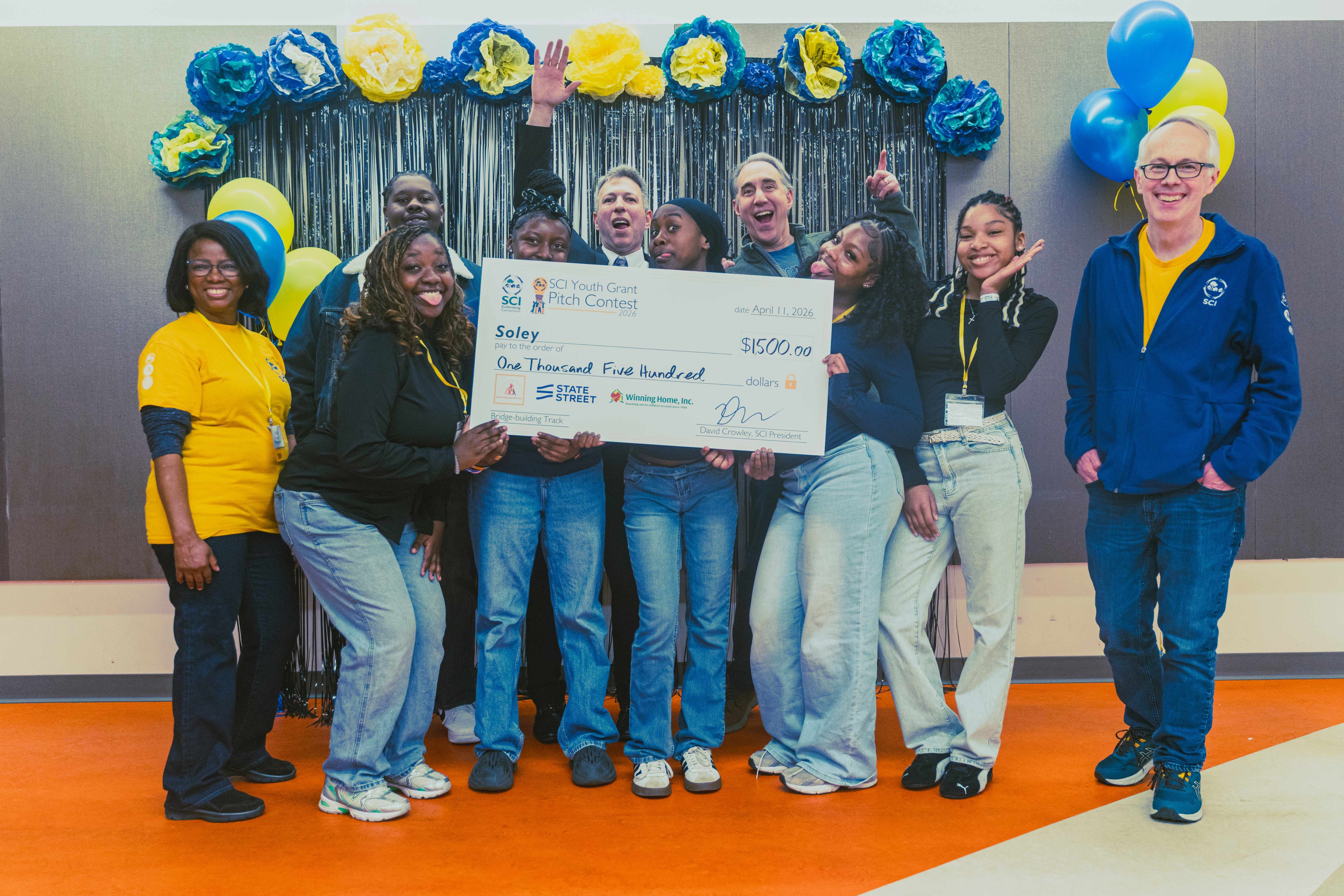 Soley's youth team members, Emalee Milcet (second on the left), Neicka Mathias, Liany Martinez, Jakyra Johnson, and Mumina Osman, pose for a fun photo with Team Advisor, Ethan Mitchell (background, left); Pitch Coaches, Jonathan Feldman (background, center) and Kenny Weill (background right); SCI Program Coordinator, Vitalie Memeus (left); and SCI President, David Crowley (right).